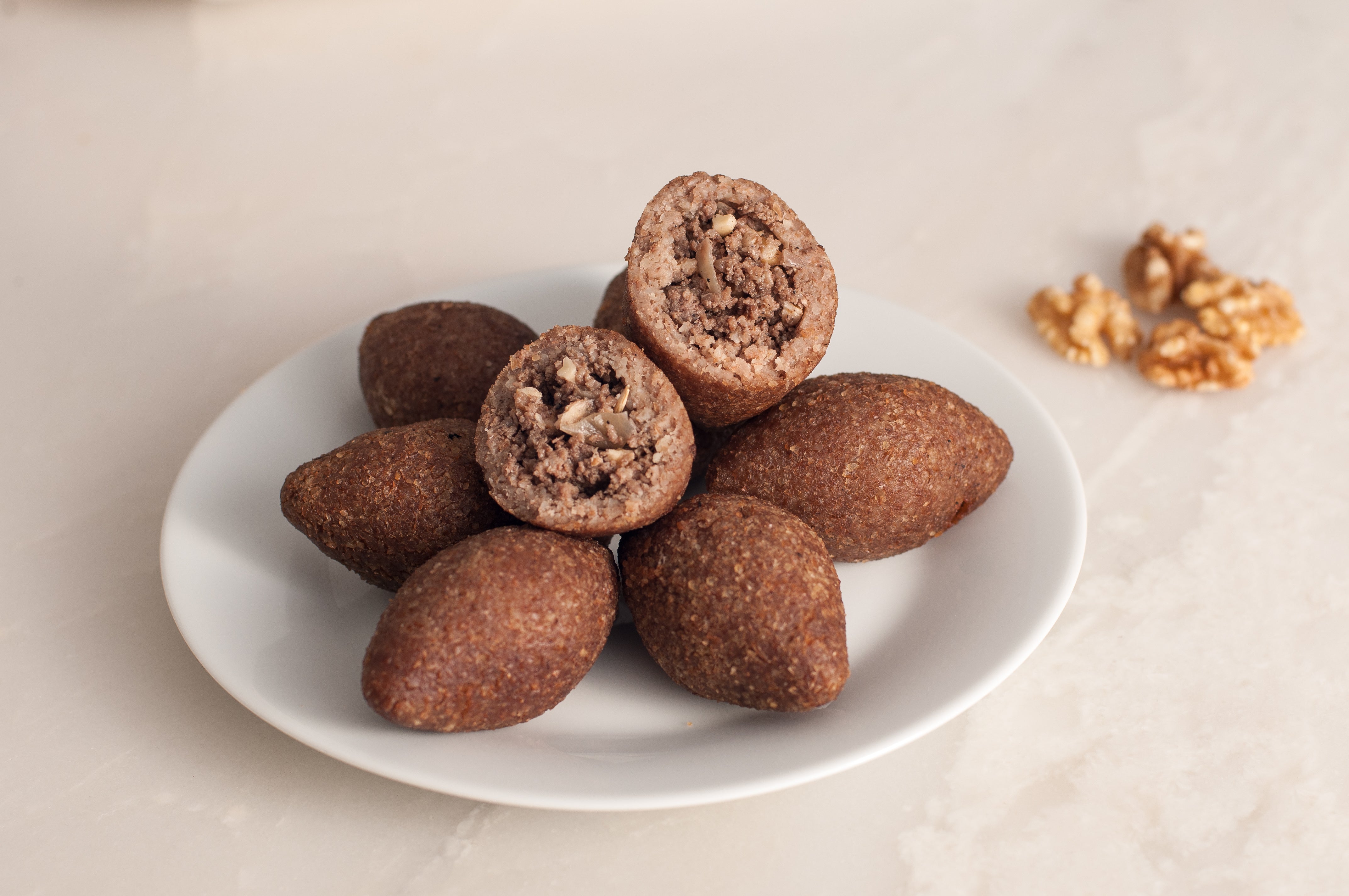 A plate of Tayybeh Kibbeh, a renowned Syrian specialty, with one cut open to reveal the savory filling infused with Middle Eastern spices, rests on a light surface. Two pieces of walnut are delicately placed on the side.