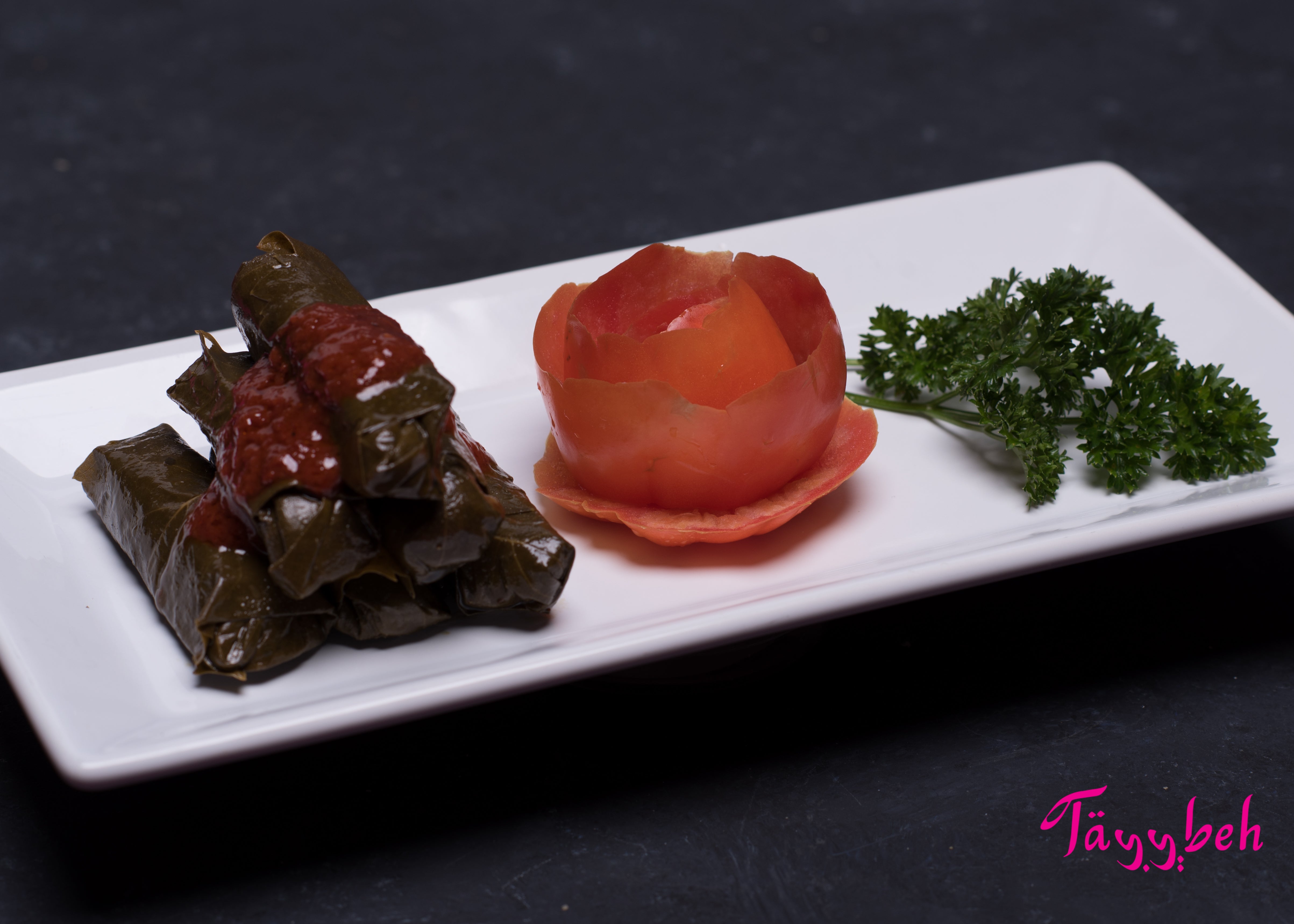 Yalanji by Tayybeh are hand-rolled vine leaves stuffed to perfection, served with a tomato garnish and a sprinkle of parsley on a rectangular white plate. These vegan delights are not only delicious but also gluten-free, offering a healthy and flavorful treat for all to enjoy.