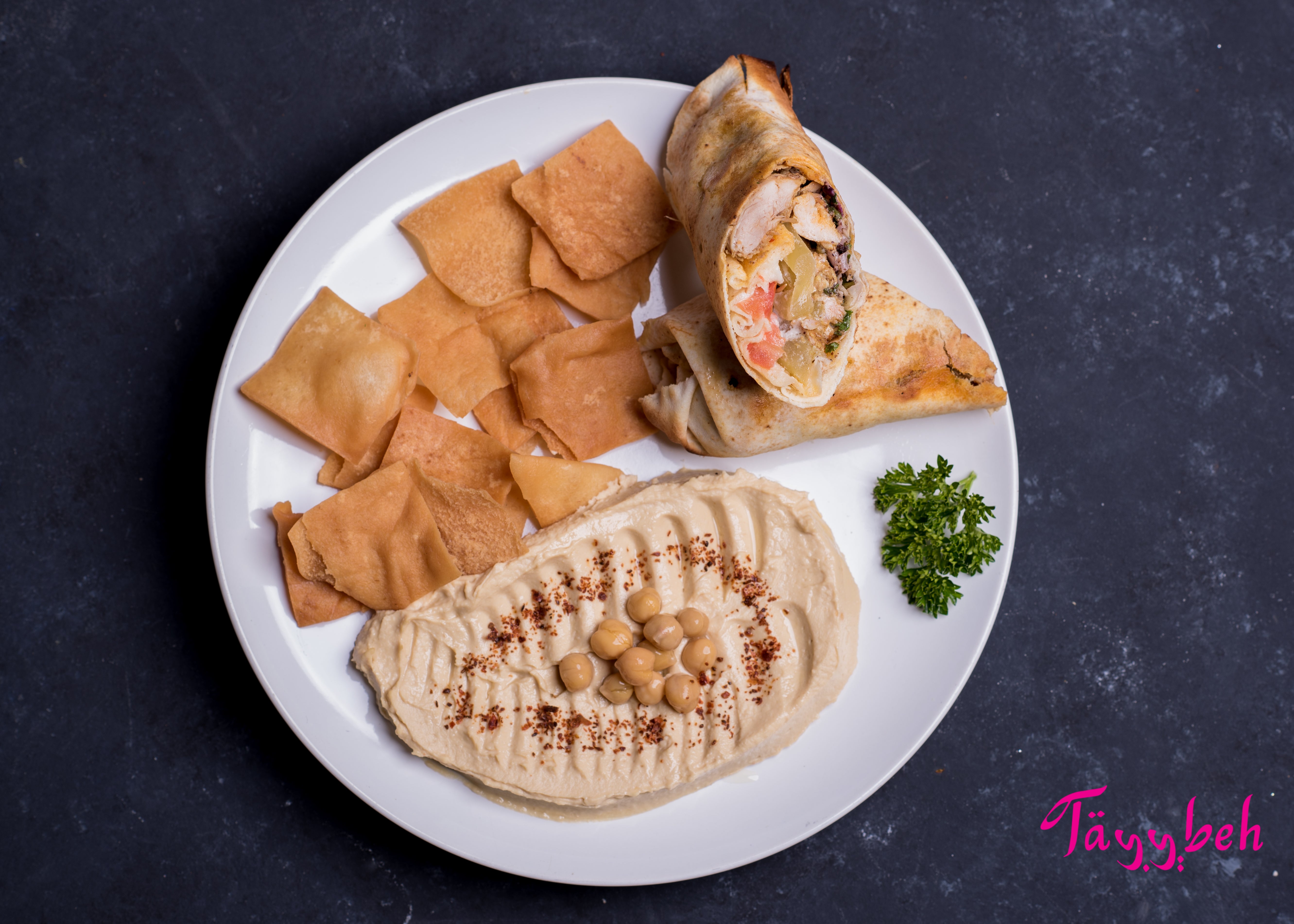 Introducing the "Chicken Shawerma Wrap Combo" by Tayybeh: A flavorful meal featuring a wrap filled with grilled chicken and vegetables, accompanied by a plate of hummus dip garnished with chickpeas and spice, served alongside sliced pita bread. Finished with a touch of parsley on the side.