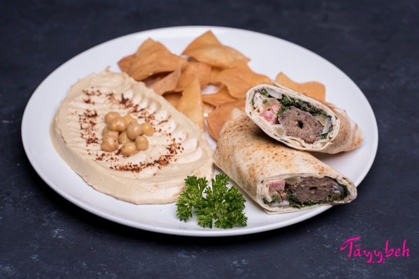 Introducing the Beef Kebab Wrap Combo by Tayybeh: Enjoy a delightful plate featuring creamy hummus dip adorned with chickpeas and spices, accompanied by crisp pita chips. Indulge in a pita wrap, halved and filled with vibrant vegetables and savory beef kebab meat, all garnished with fresh parsley.