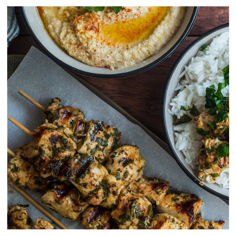 The Latakia, from Tayybeh, is beautifully arranged on a wooden table with grilled chicken skewers, a bowl of hummus with paprika, and a plate of rice and vermicelli garnished with herbs.