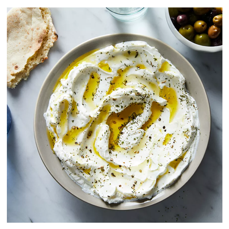 A bowl of Tayybeh Labneh dip drizzled with olive oil and garnished with mint sits on a marble surface, alongside a piece of bread and a bowl of olives.