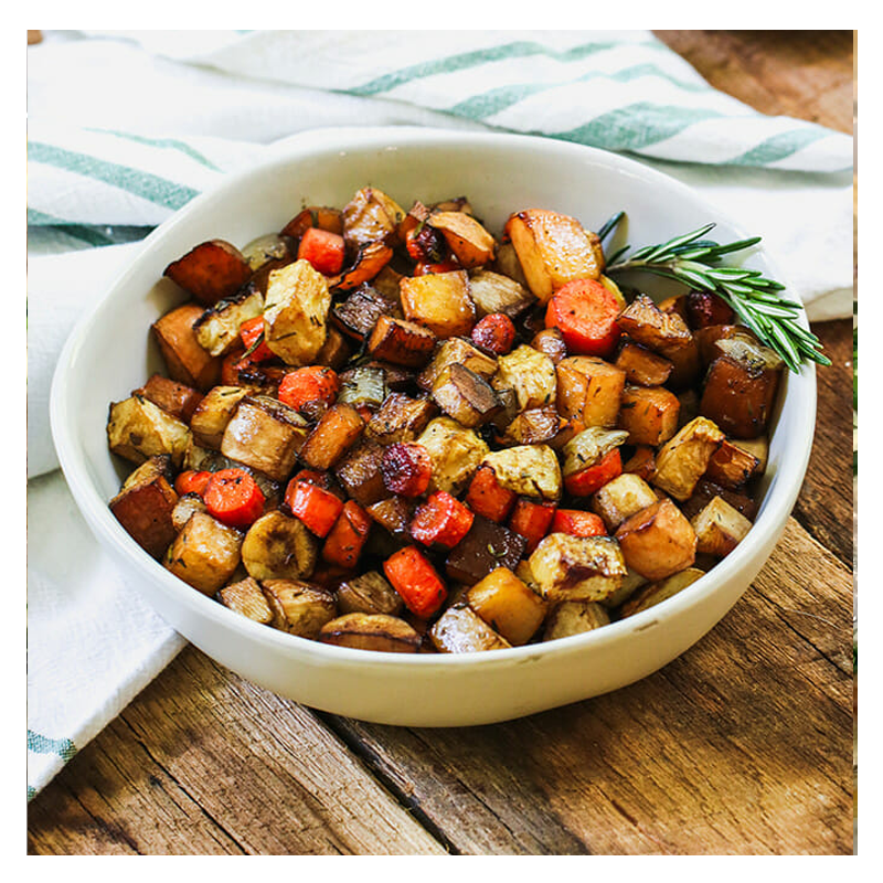 A Tayybeh "Roast Vegetables" bowl featuring oven-roasted root vegetables like carrots and potatoes, garnished with a sprig of rosemary, sits on a wooden table with a striped cloth.