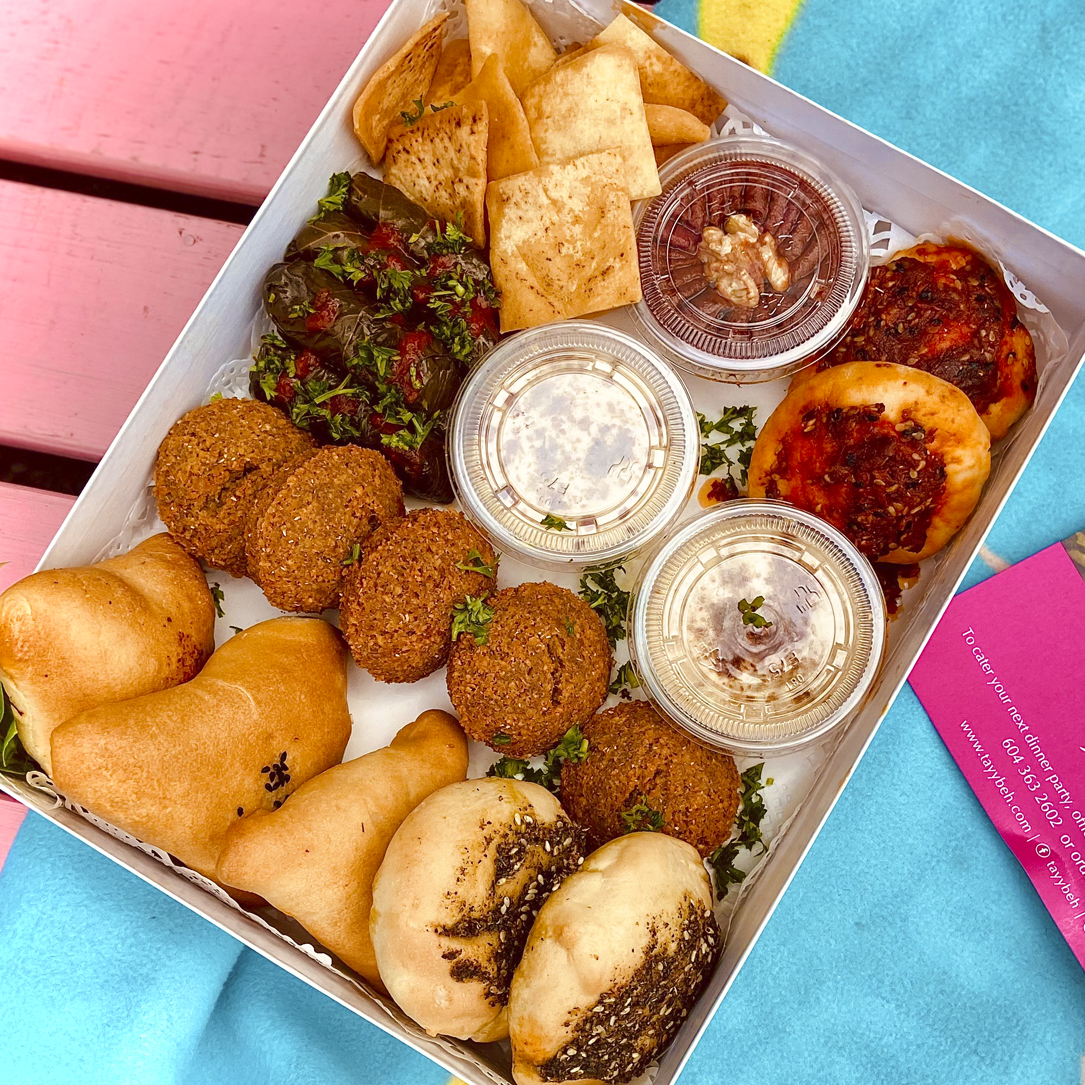 A Sampler Box for two by Tayybeh, featuring a selection of Middle Eastern appetizers such as vegan falafel, stuffed grape leaves, assorted flatbreads with toppings, gluten-free pita chips, and various dips in small containers.