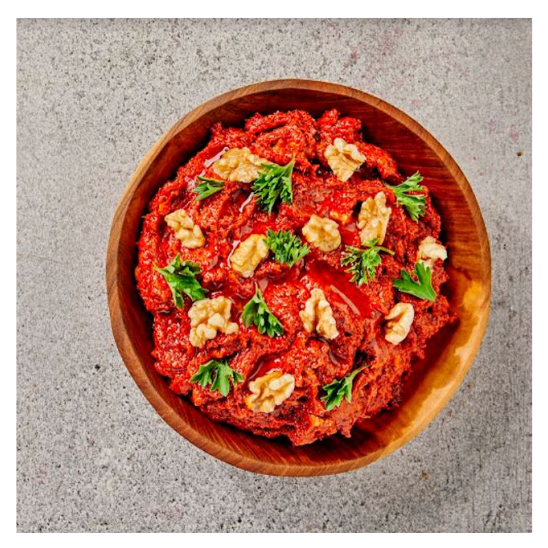 A wooden bowl filled with Tayybeh's Mhammara dip, topped with walnuts and parsley, is set against a gray background.