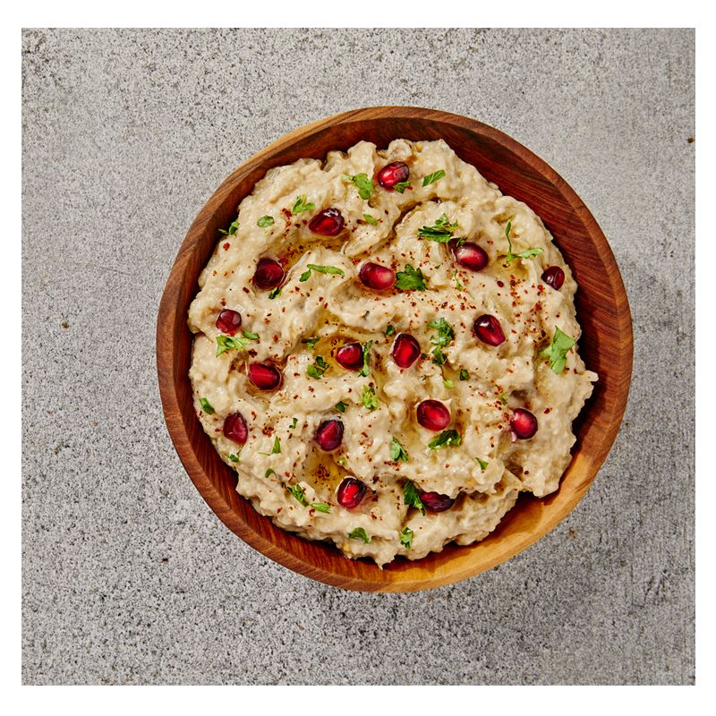 A wooden bowl of Tayybeh's Mutabbal Dip, swirled with tahini and topped with pomegranate seeds and fresh herbs, is placed on a textured gray surface. Serve it alongside crispy pita chips for a delightful crunch.