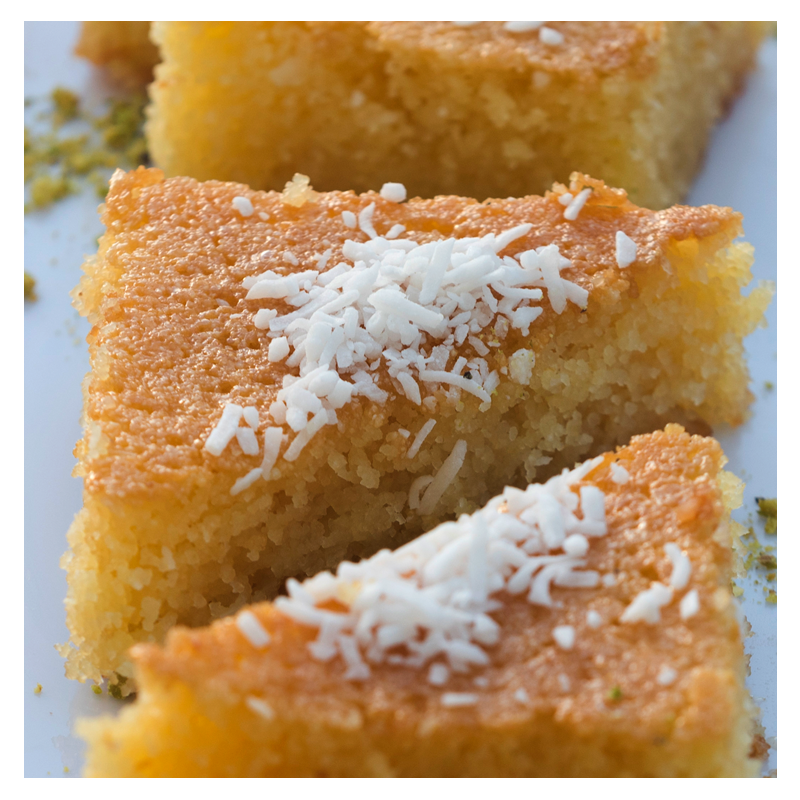 A close-up shot of three triangular servings of Tayybeh's Nammoura, a golden and moist semolina cake topped with coconut flakes, arranged on a white plate for an irresistible sweet experience.