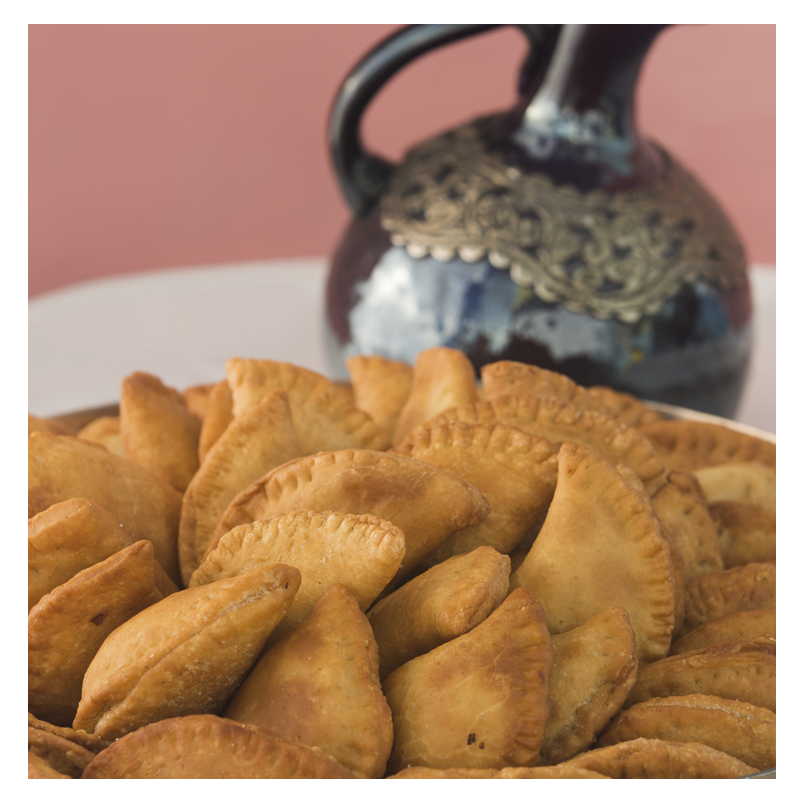 In the foreground is a plate of Golden Crispy half-moon pockets stuffed with beef and parsley, known as Tayybeh's Frozen Beef Samboosak (dozen), while an ornately designed dark ceramic jug adorns the blurred background. These enticing samboosaks are completely nut-free and dairy-free, providing a delightful appetizer for everyone to enjoy.