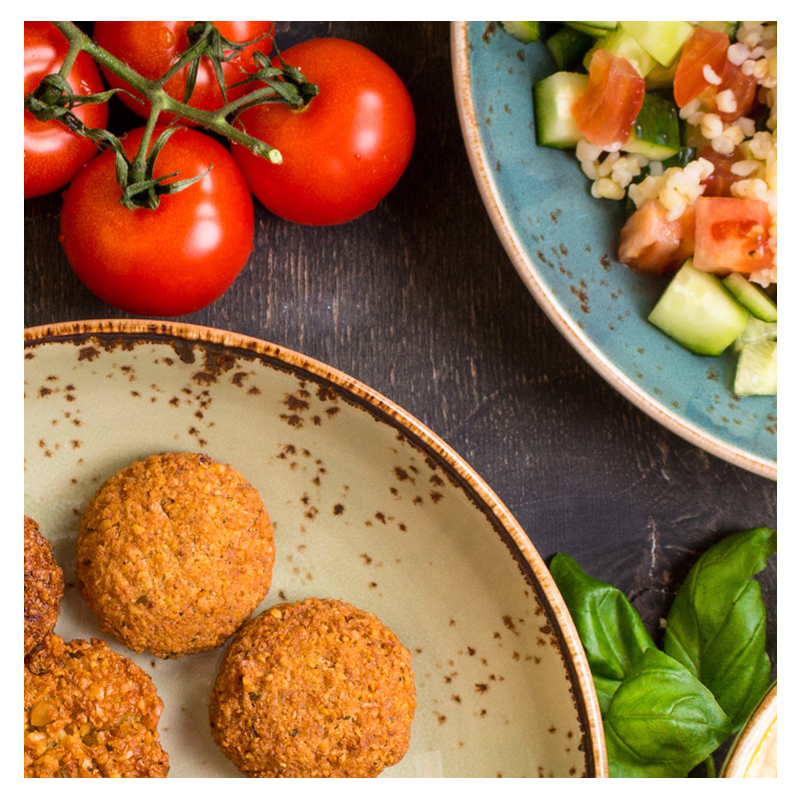 The Vegan plate by Tayybeh features falafel, fresh vine tomatoes, and a bowl of bulgur salad with cucumber and tomato, all perfectly complemented by creamy hummus on a dark surface.