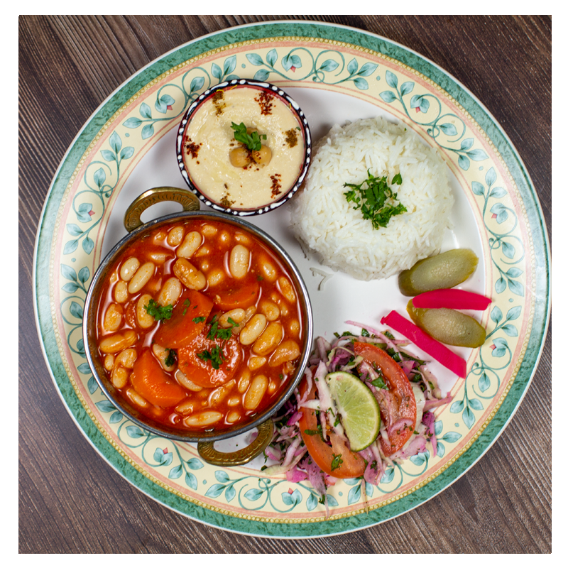 A serving of fluffy white rice paired with Tayybeh's White Bean and Carrot Stew Meal, alongside hummus with toppings, a tomato and onion salad dressed with lime, and pickled vegetables on a decorative plate. Each dish is carefully prepared to ensure it remains gluten-free.