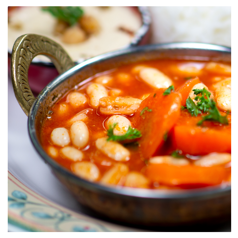 A close-up of Tayybeh's White Bean and Carrot Stew Meal reveals a gluten-free dish loaded with carrots and garnished with herbs. In the background, a second dish featuring rice is blurred, making it an ideal vegan pairing.