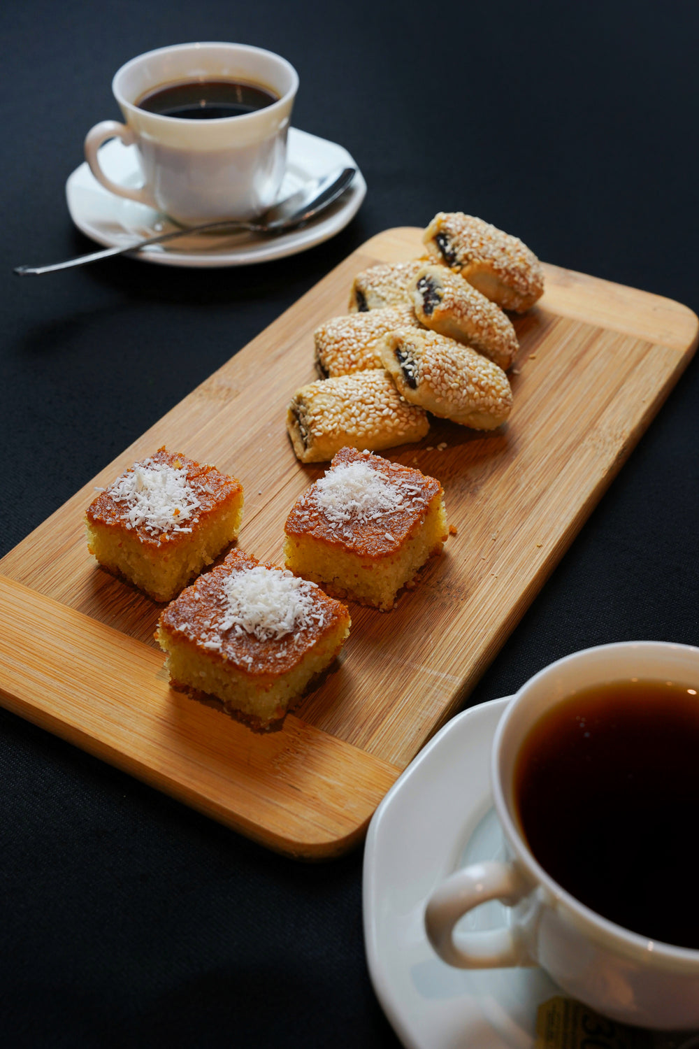 The "Coffee Break Package 2" by Tayybeh, featuring a wooden tray with two cups of fair-trade coffee, coconut-topped nammoura squares, and sesame-seed-covered barazek pastries, rests elegantly on a black table.