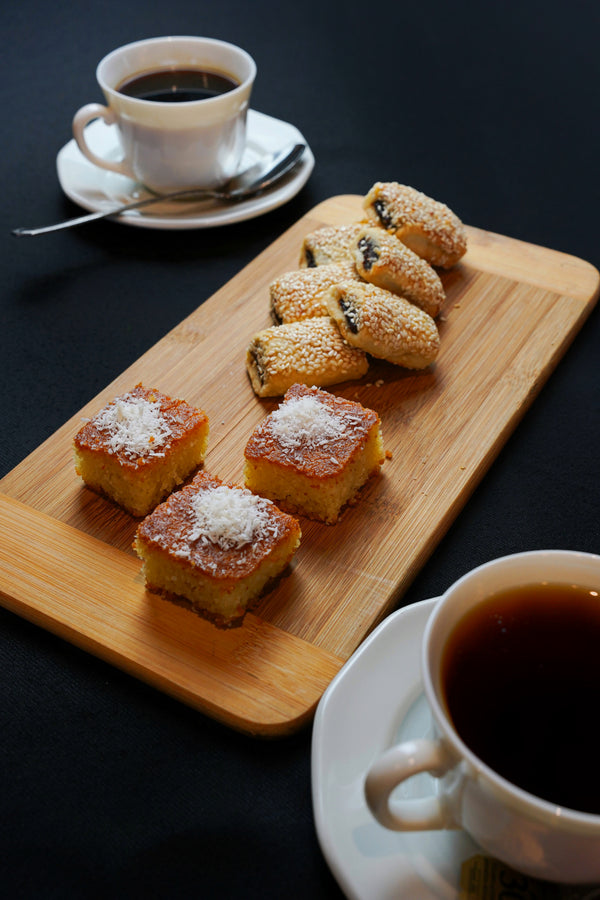 The "Coffee Break Package 2" by Tayybeh, featuring a wooden tray with two cups of fair-trade coffee, coconut-topped nammoura squares, and sesame-seed-covered barazek pastries, rests elegantly on a black table.