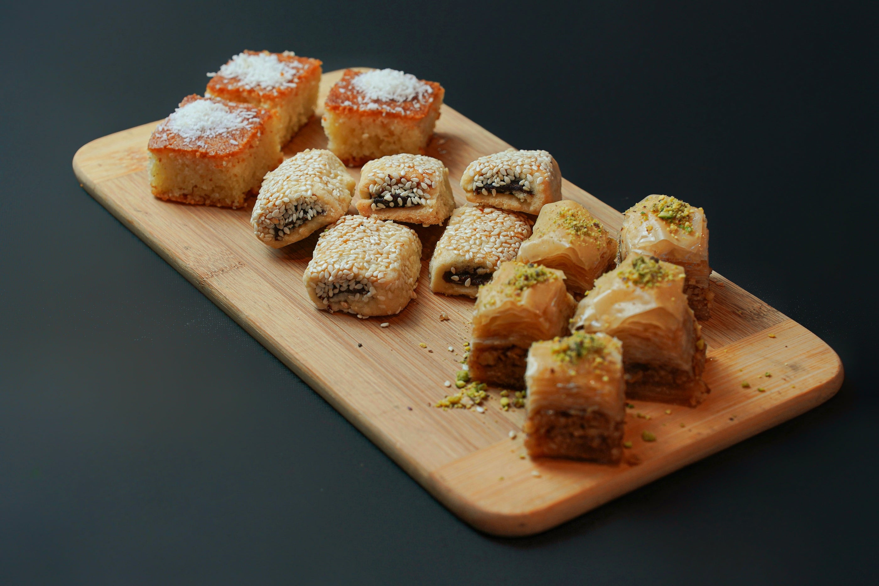 A wooden board featuring the Assorted Dessert Platter by Tayybeh, which includes coconut-topped Nammoura squares, sesame-covered pastry rolls, and pistachio-topped baklava.