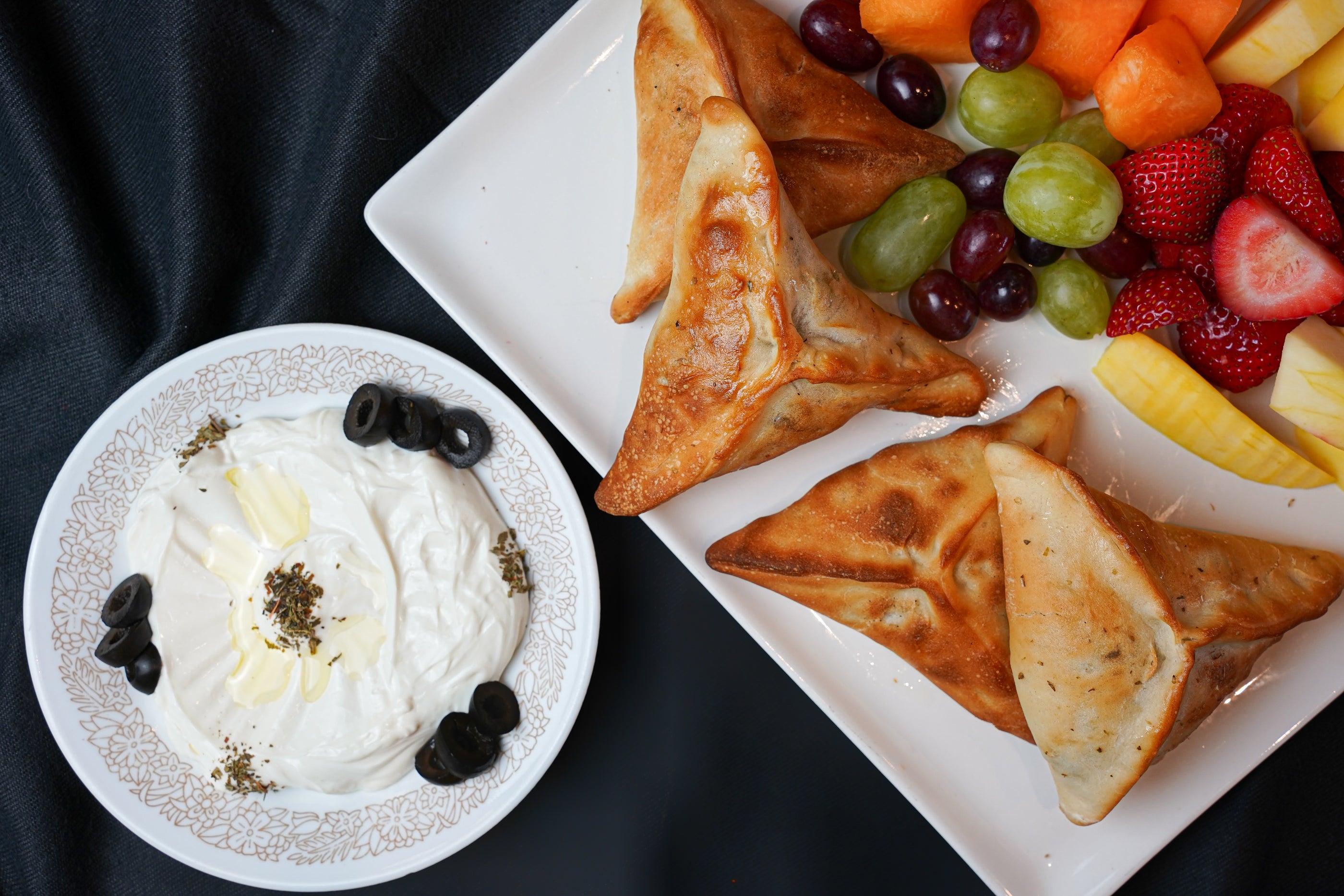 A plate with samosas and a vibrant fruit platter sits next to a bowl of labneh dip, topped with black olives and herbs, as part of the Sunrise Breakfast from Tayybeh.