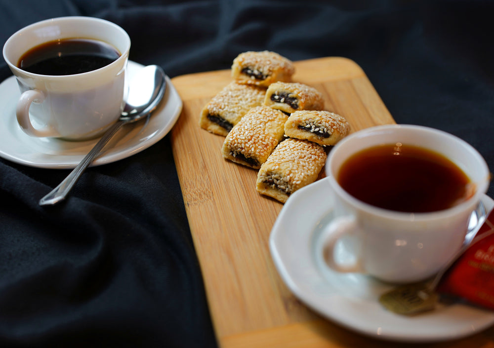 Tayybeh's Coffee Break Package 1 includes two cups of tea on saucers with spoons, accompanied by a wooden board featuring date cookies and sesame pastries, all elegantly arranged on a black tablecloth.