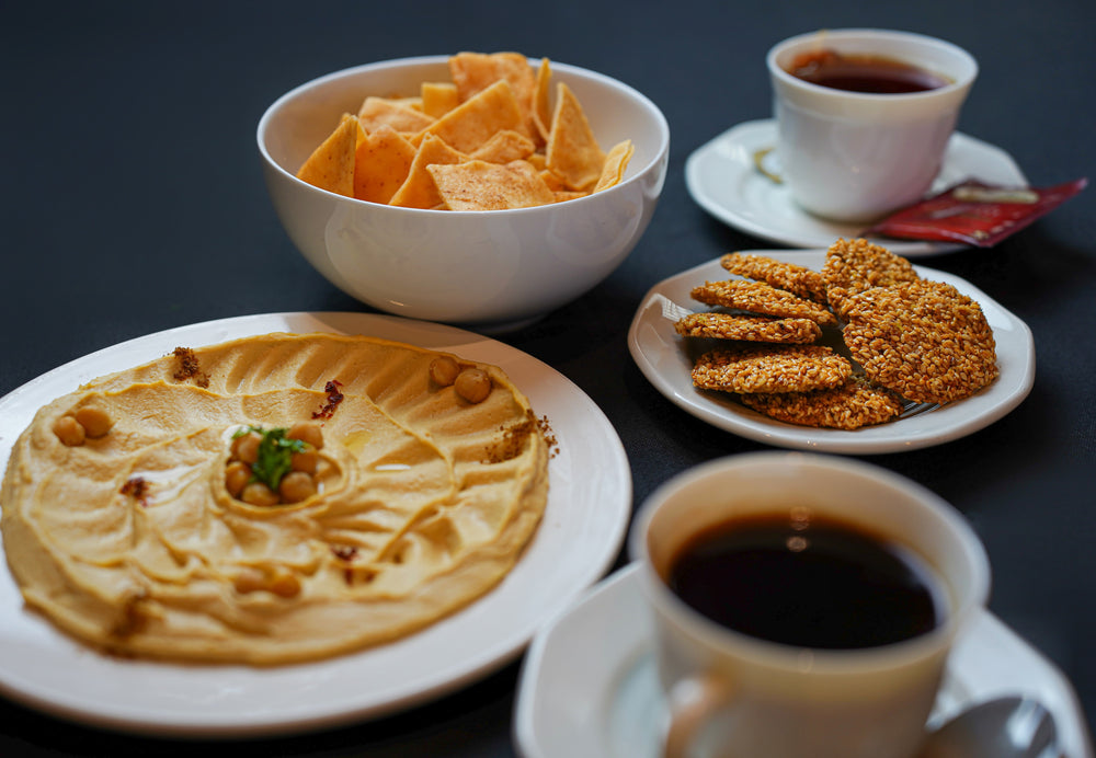 A table featuring Tayybeh's Coffee Break Package 3, which includes hummus dip garnished with chickpeas and herbs, a bowl of pita chips, sesame seed cookies, a slice of date and walnut cake, accompanied by two cups of tea on saucers.