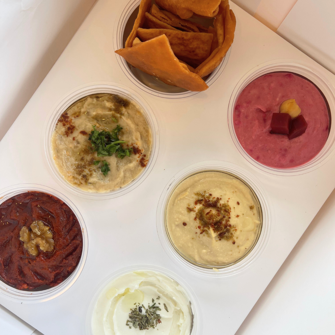 The Beach Box by Tayybeh features a selection of six small containers filled with vibrant dips such as beet, yogurt, and hummus, complemented by crispy pita chips, all elegantly arranged on a white surface.