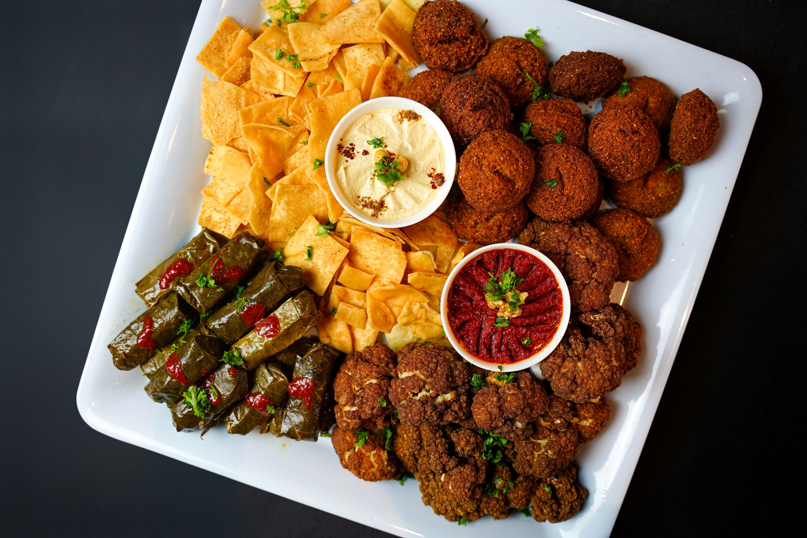 Introducing the Tayybeh Mixed Appetizer Platter: a white square plate featuring falafel, grape leaves, crispy pita chips, and two small bowls of hummus dip—a perfect vegan delight.