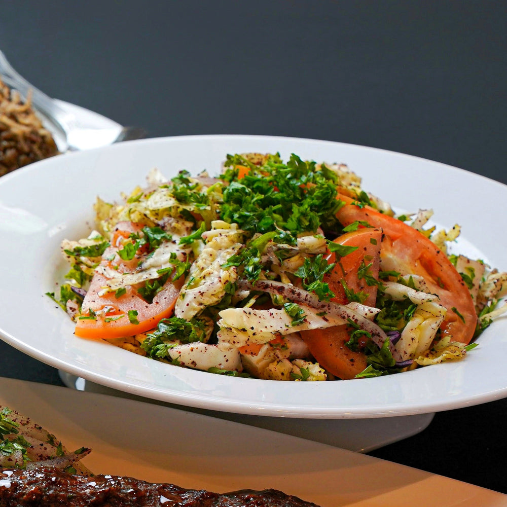 A bowl of Tayybeh's Cabbage Salad, featuring fresh tomatoes, lettuce, parsley, and crunchy cabbage, displayed on a dark tabletop.