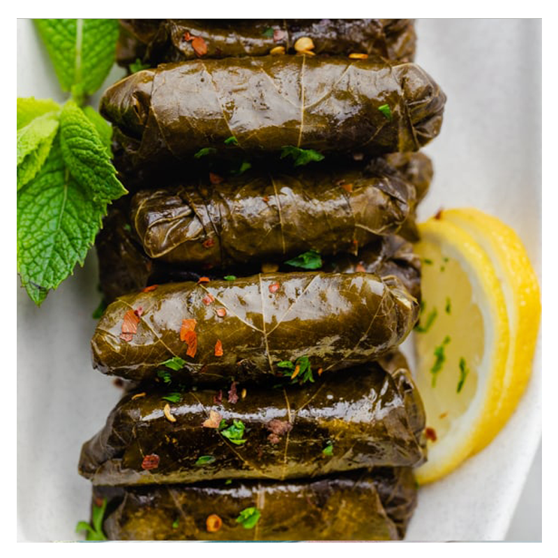 On a white plate, a stack of Tayybeh's Yalanji, stuffed vine leaves filled with aromatic rice and seasoned with herbs and spices, is garnished with mint leaves and a lemon slice.