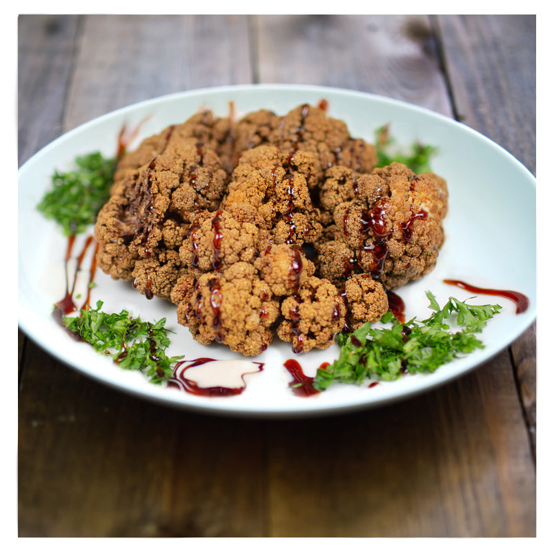 Tayybeh's Crispy Cauliflower, a dish of golden florets deep-fried to perfection and enhanced with pomegranate molasses, is elegantly garnished with fresh parsley on a wooden table.